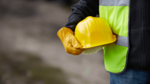 Image shows a tradesman holding his yellow hard hat wear high vis vest and yellow glove,