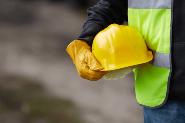 Image shows a tradesman holding his yellow hard hat wear high vis vest and yellow glove, to represent working on Social Housing Construction project