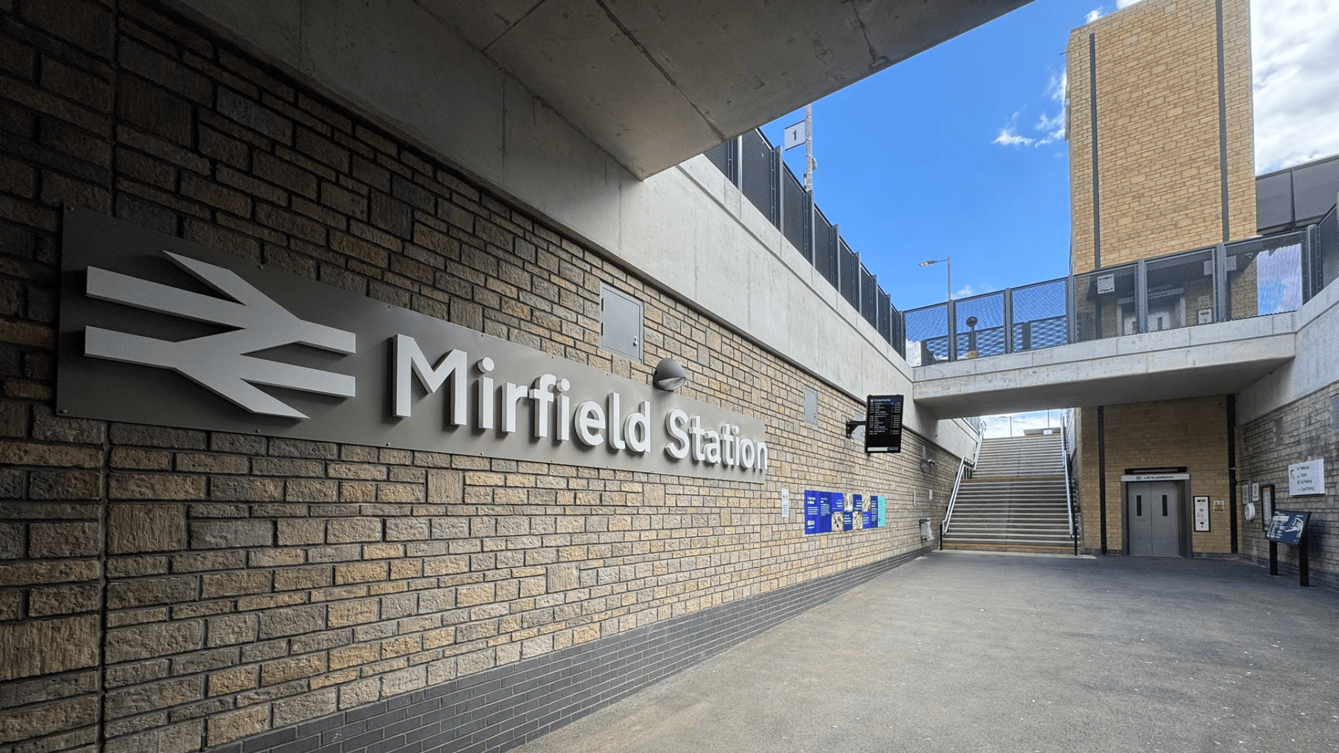 A covered station entrance area featuring a large Mirfield Station sign mounted on a brick wall. Wide steps lead up to the platforms, while a lift provides step free access on the right. Information boards, digital displays, and wayfinding signage are positioned throughout the space, with modern brickwork, concrete structures, and open access routes creating a bright, accessible station environment.