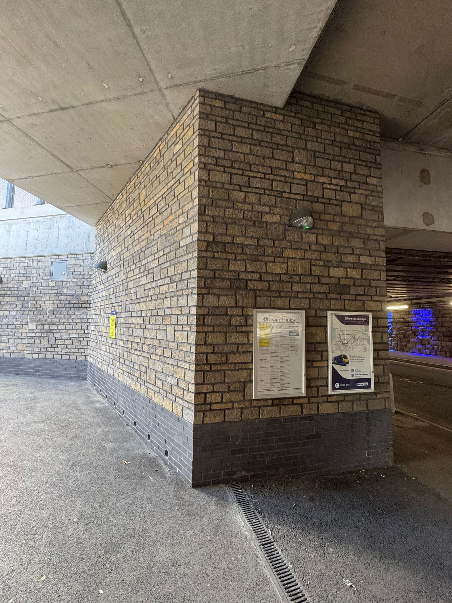 A covered station walkway with brick clad walls, concrete ceilings, and wall mounted lighting. Information boards displaying train times and travel information are fixed to a brick pillar, while a drainage channel runs along the ground. The space connects to an underpass area illuminated with blue accent lighting in the distance.