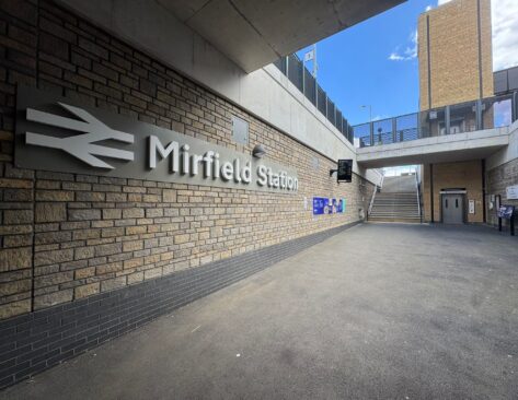 A covered station entrance area featuring a large Mirfield Station sign mounted on a brick wall. Wide steps lead up to the platforms, while a lift provides step free access on the right. Information boards, digital displays, and wayfinding signage are positioned throughout the space, with modern brickwork, concrete structures, and open access routes creating a bright, accessible station environment.