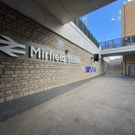 A covered station entrance area featuring a large Mirfield Station sign mounted on a brick wall. Wide steps lead up to the platforms, while a lift provides step free access on the right. Information boards, digital displays, and wayfinding signage are positioned throughout the space, with modern brickwork, concrete structures, and open access routes creating a bright, accessible station environment.