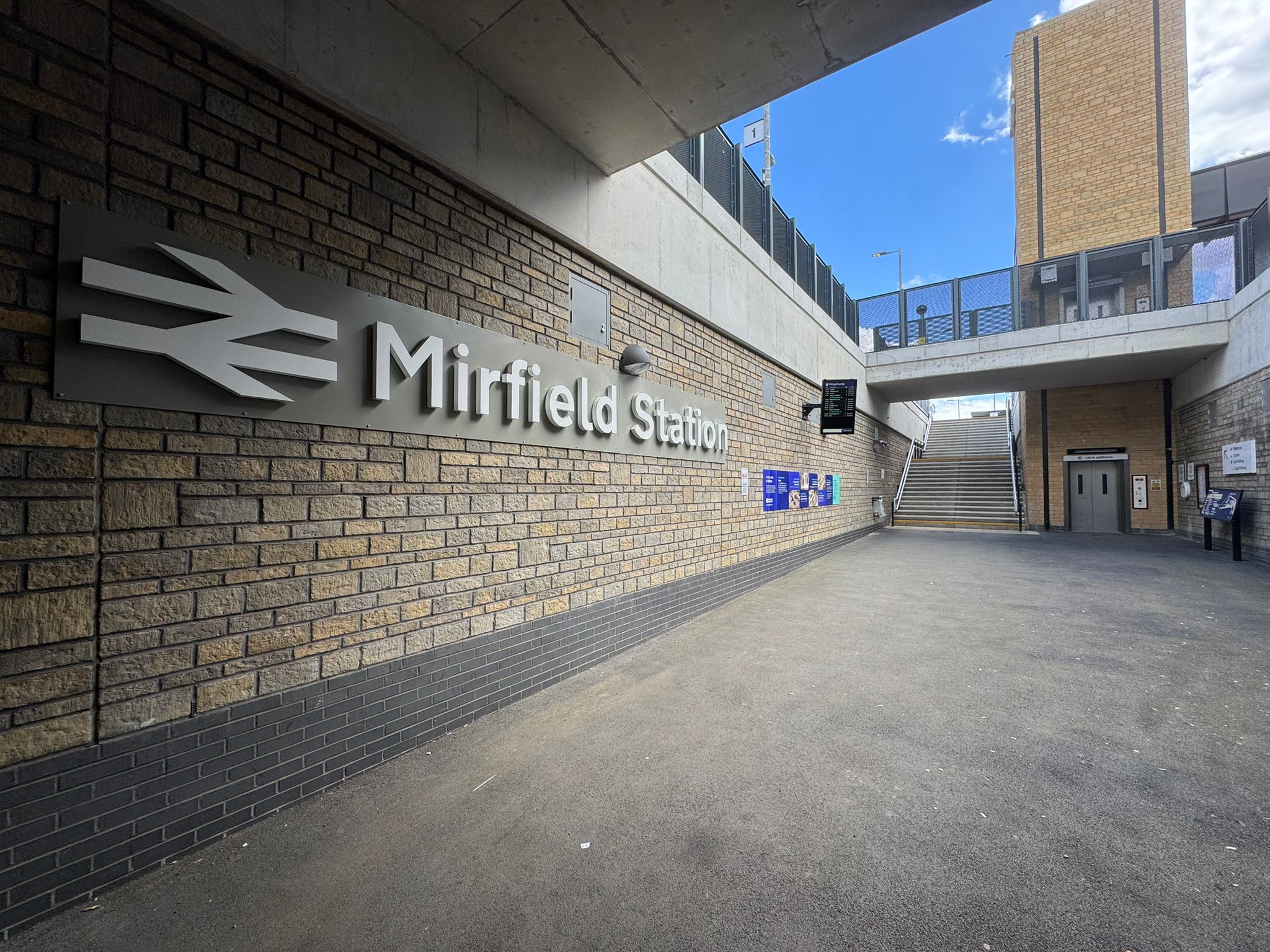 A covered station entrance area featuring a large Mirfield Station sign mounted on a brick wall. Wide steps lead up to the platforms, while a lift provides step free access on the right. Information boards, digital displays, and wayfinding signage are positioned throughout the space, with modern brickwork, concrete structures, and open access routes creating a bright, accessible station environment.