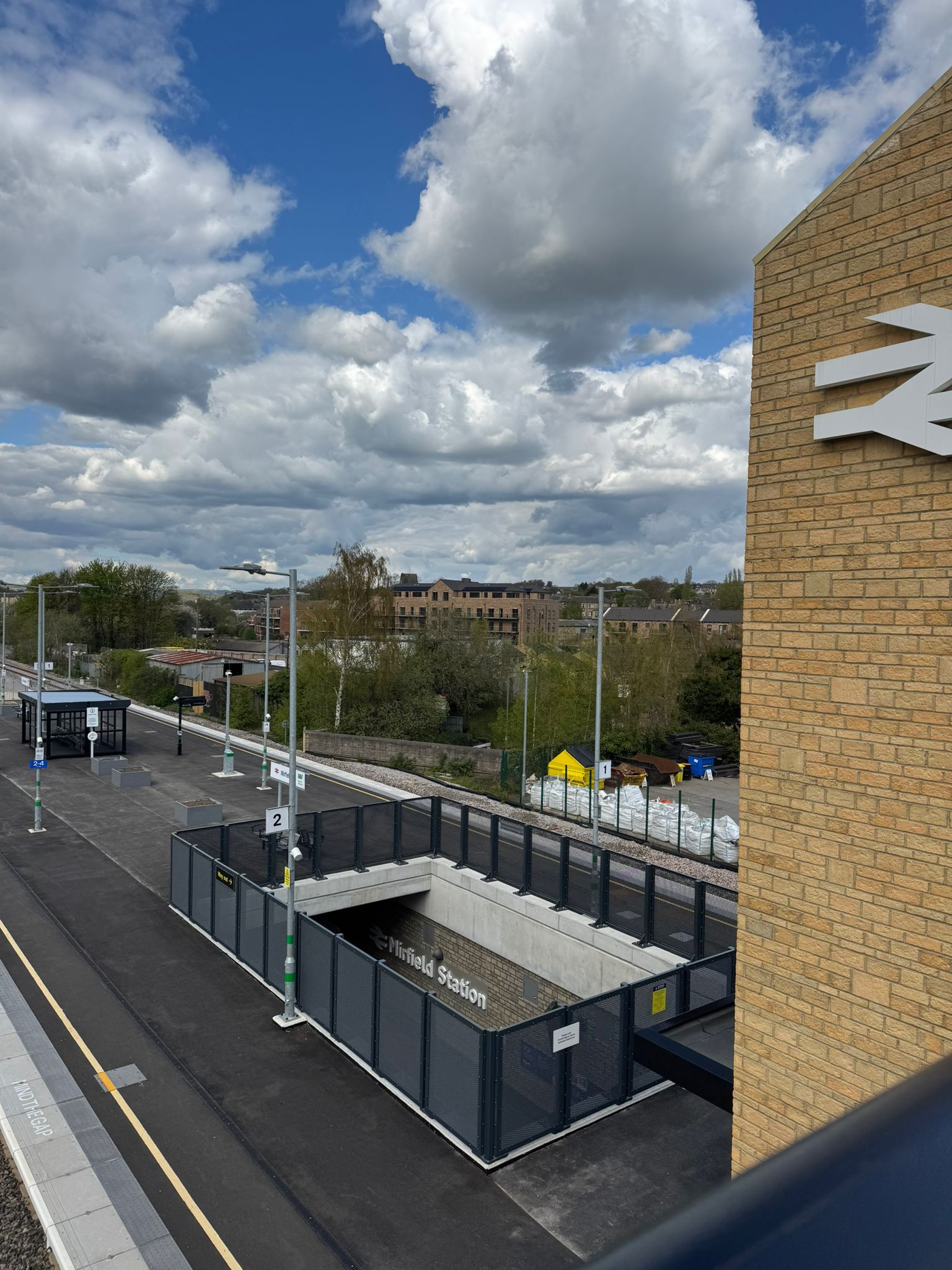 Elevated view across Mirfield Station showing platform areas, safety fencing, lighting columns, and station signage. A brick clad station tower with a partial rail symbol is visible in the foreground, while the station entrance sits below within a fenced structure. Trees, buildings, and a dramatic cloud filled sky form the backdrop.