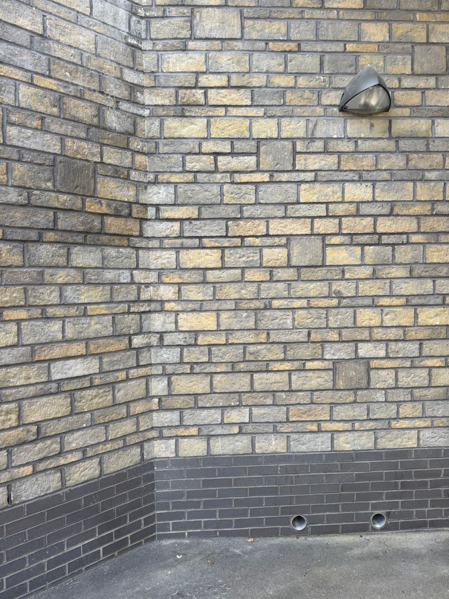 Close up of a curved brick wall within a station underpass, showcasing detailed masonry work in mixed buff and grey tones. A wall mounted light fitting is positioned above, while dark brick detailing run along the base of the structure. The image focuses on the construction finish and brickwork craftsmanship.