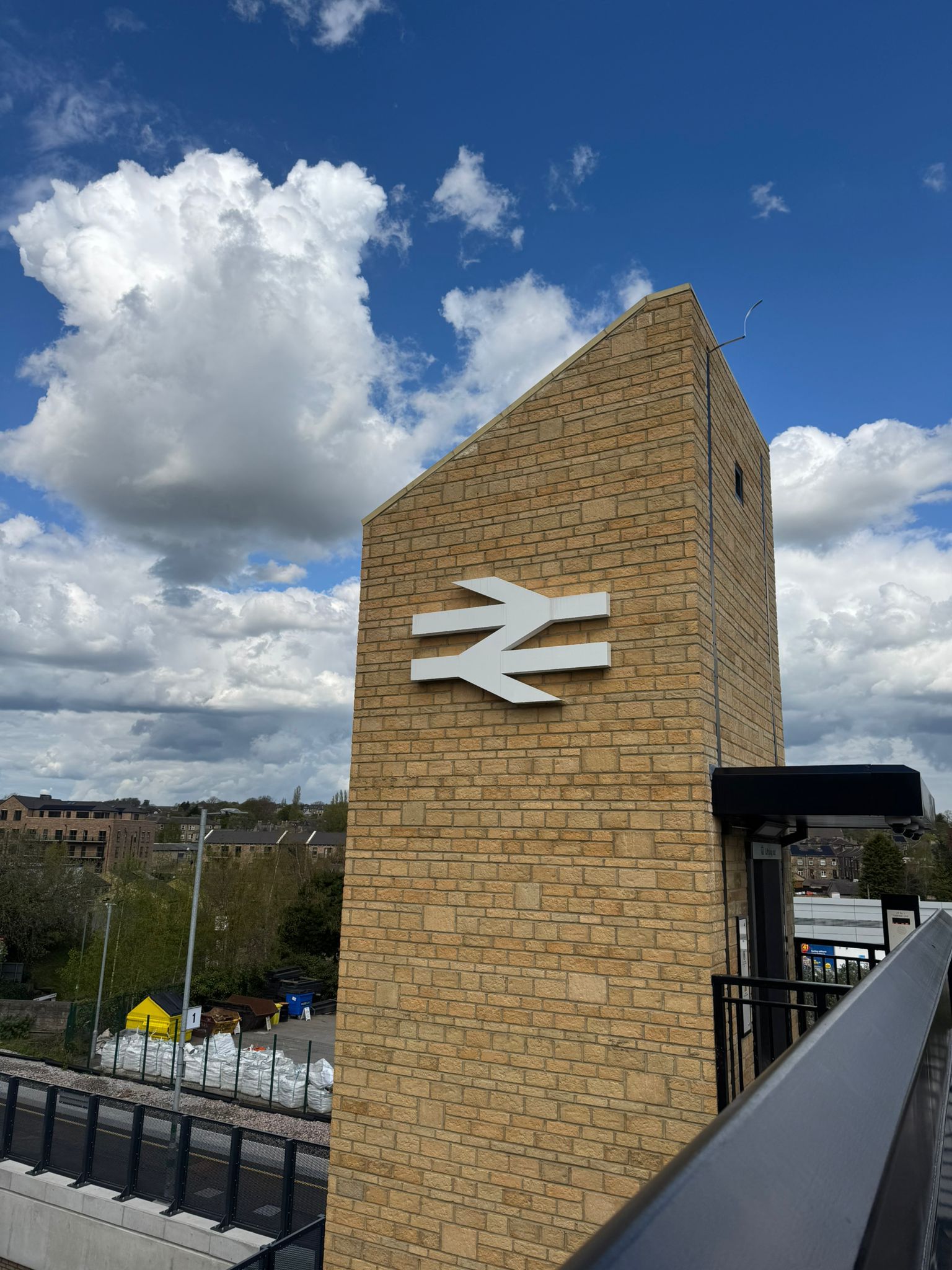 A brick clad station tower displaying a large white rail symbol stands against a bright blue sky with large white clouds. The image is taken from an elevated walkway with black railings, overlooking parts of the station, surrounding buildings, and platform infrastructure below.
