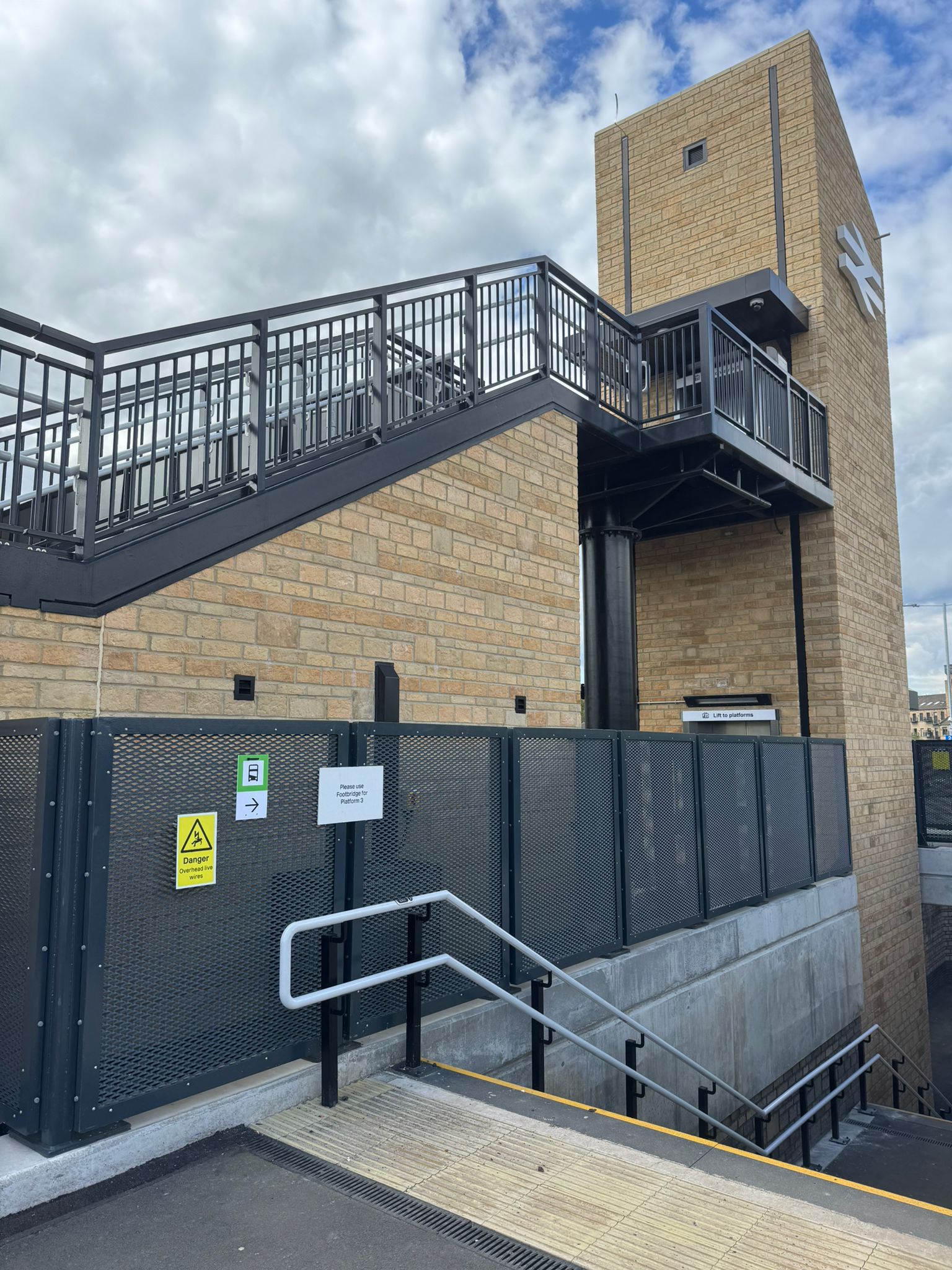 A brick clad station structure featuring a staircase, lift tower, and elevated walkway with black steel railings. In the foreground, steps with handrails lead down to a lower level, while metal mesh safety fencing surrounds the platform edge. Directional and safety signs are mounted on the fencing, with a partly cloudy sky overhead.