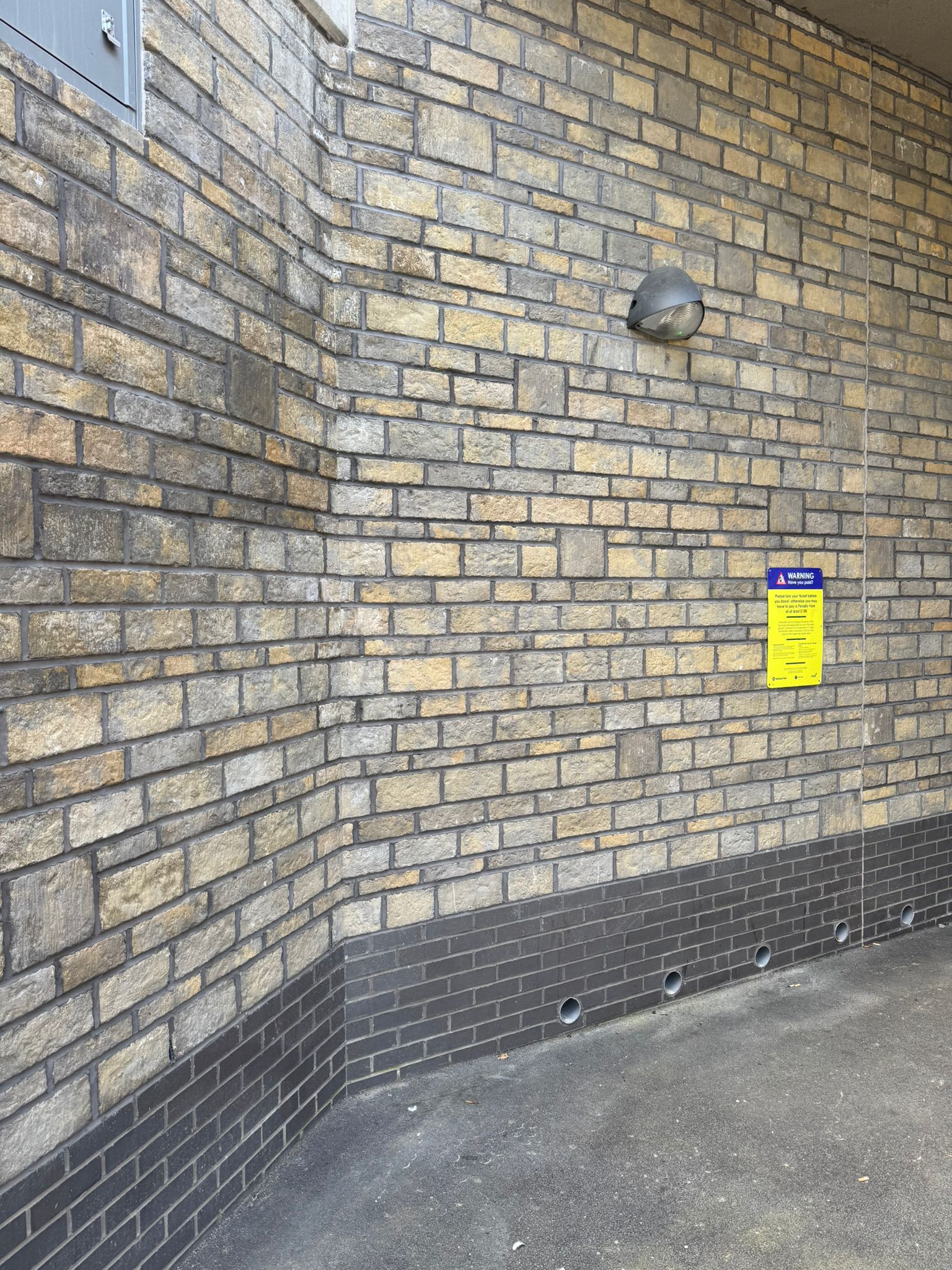 Close up of a curved brick wall within a covered station area, featuring mixed tone brickwork, a wall mounted light, ventilation openings at ground level, and a yellow safety notice. The image highlights the construction detailing and masonry finish of the station structure.