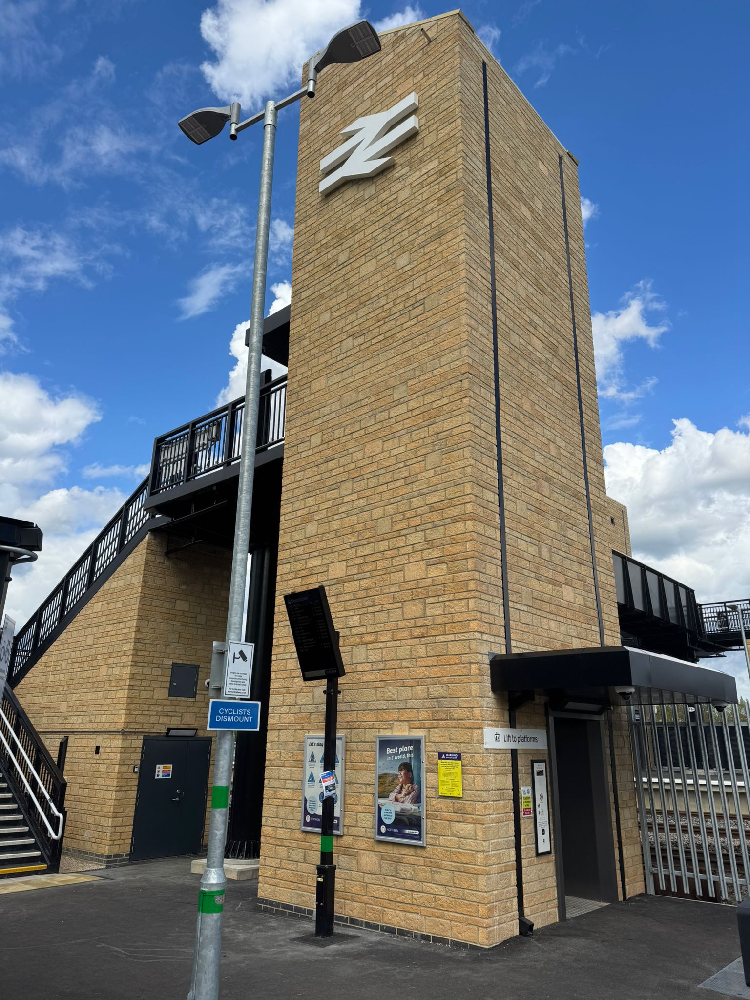 A tall brick clad lift tower and adjoining staircase structure at Mirfield railway station. The tower features a large white rail symbol near the top, with lift access at ground level beneath a dark canopy. A staircase leads to an elevated footbridge, while lighting columns, information boards, and safety signage are positioned around the station forecourt. A bright blue sky with scattered clouds forms the backdrop.
