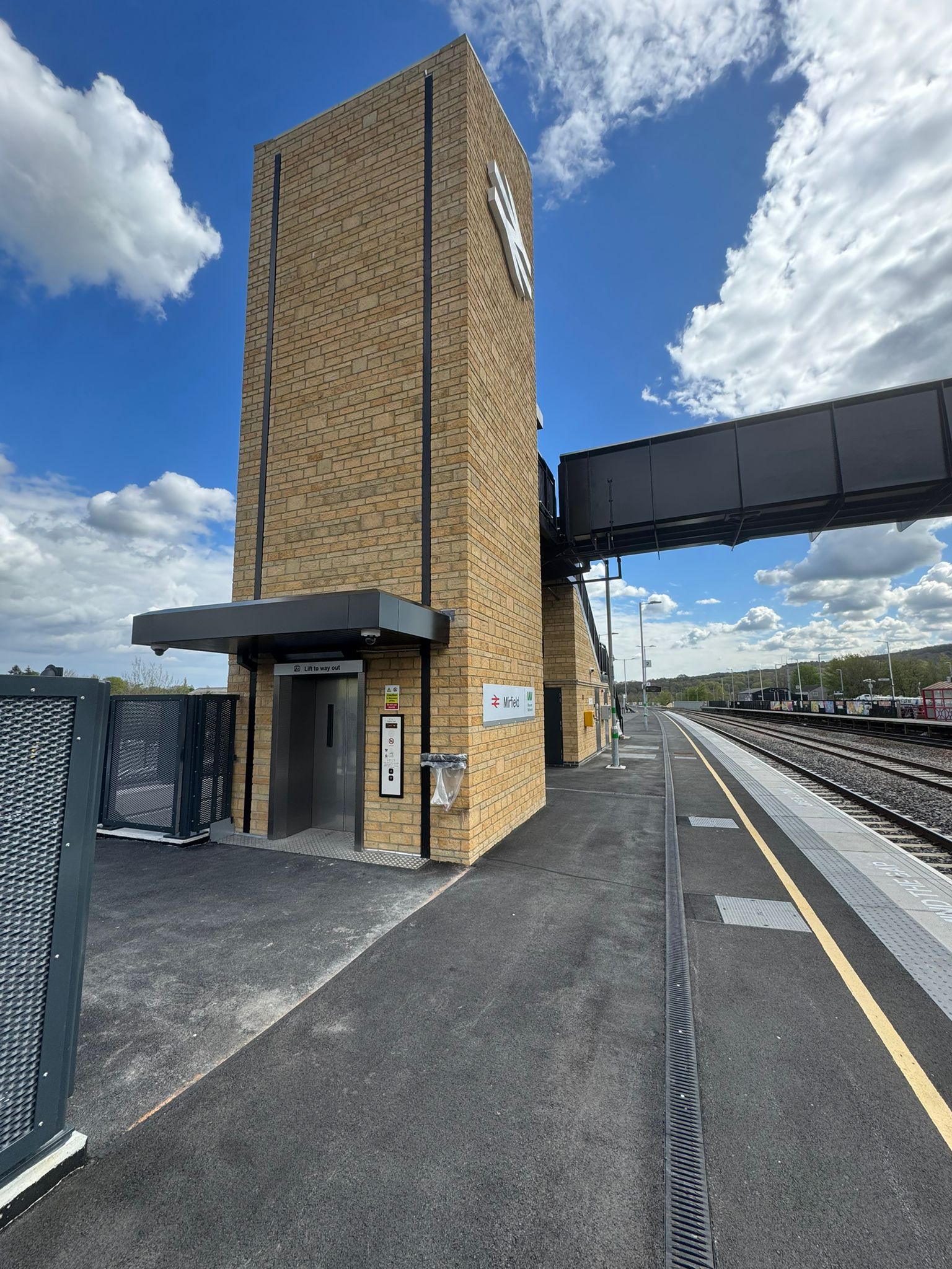 A brick clad lift tower stands beside a railway platform, connected to an elevated pedestrian footbridge. The tower features a lift entrance beneath a dark canopy, with platform signage and safety markings visible nearby. Railway tracks run alongside the platform, with open views of the station and a bright sky filled with large white clouds.