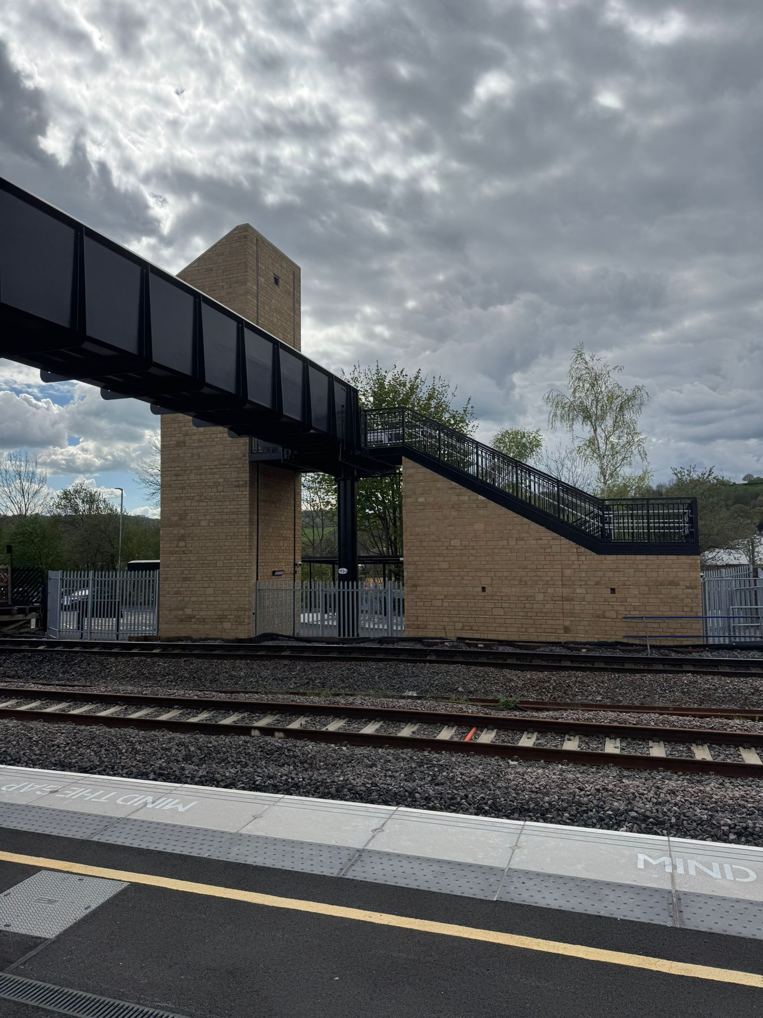 View across railway tracks towards a modern pedestrian footbridge and staircase structure at Mirfield railway station. The bridge features black steel framing, brick clad support towers, and stair access between platform levels. Trees and fencing surround the station, with dramatic grey clouds overhead.
