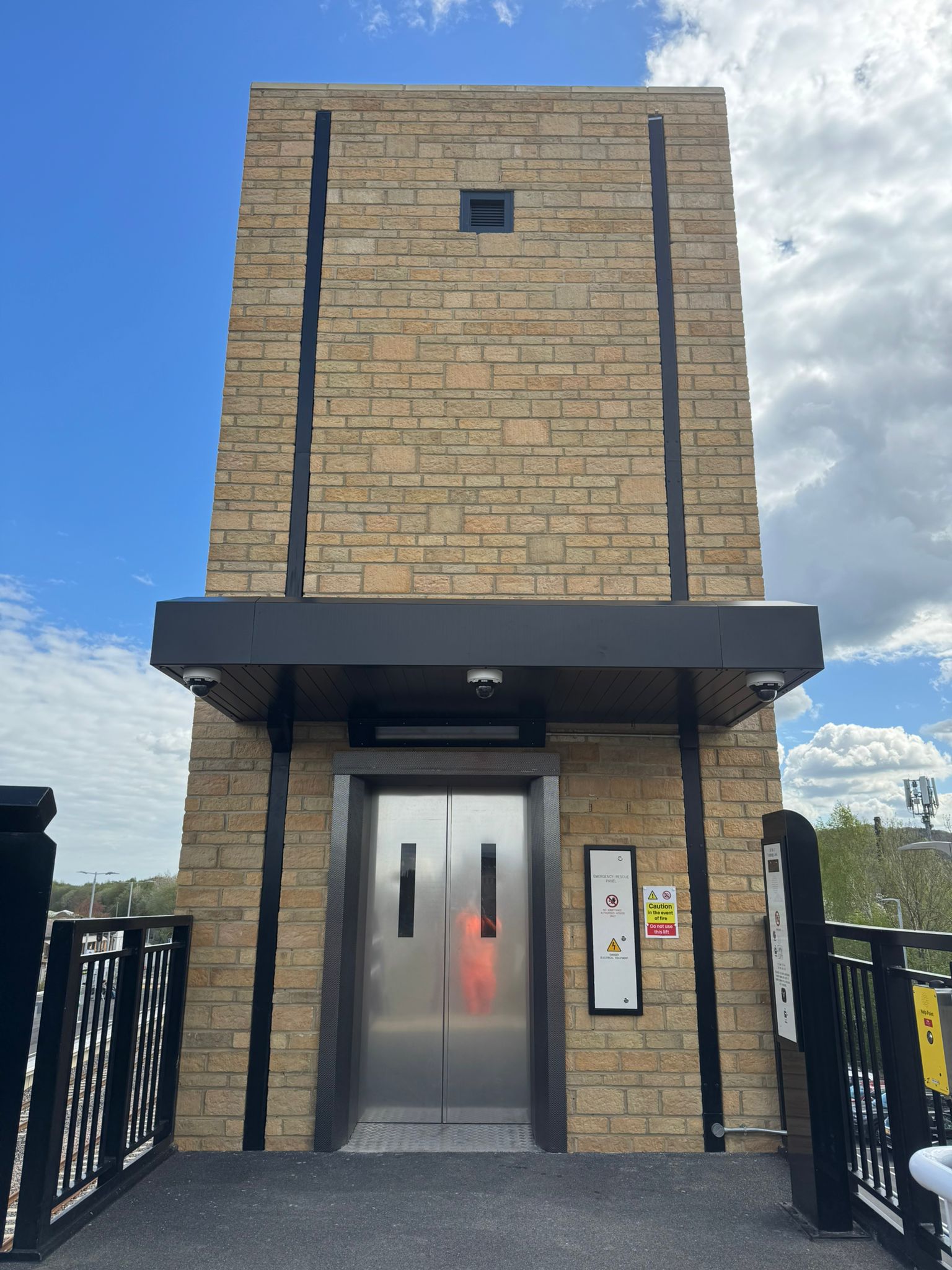 A tall stone clad lift tower with stainless steel lift doors stands on a railway platform. Black steel railings line the platform edge, and modern signage is mounted beside the lift entrance. The structure features a dark canopy above the doors and security cameras underneath. Blue sky and clouds form the background, highlighting the newly constructed station infrastructure.