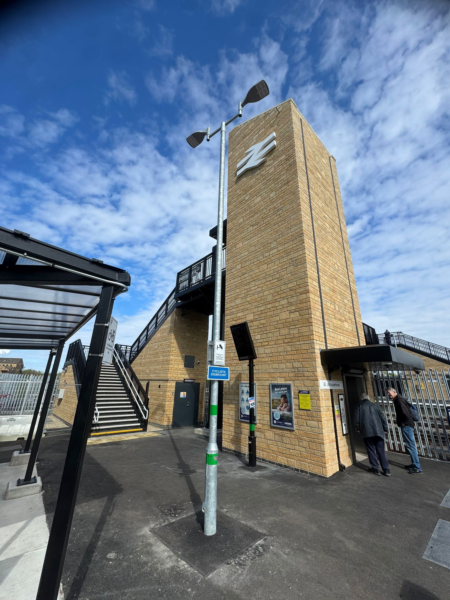 A brick clad station tower displaying a large white rail symbol stands beside a staircase and lift entrance. Black steel railings, platform shelters, information boards, and lighting columns surround the structure. Two people stand near the lift entrance, while a bright blue sky with scattered clouds forms the backdrop. The image showcases the station's modern access and circulation facilities.