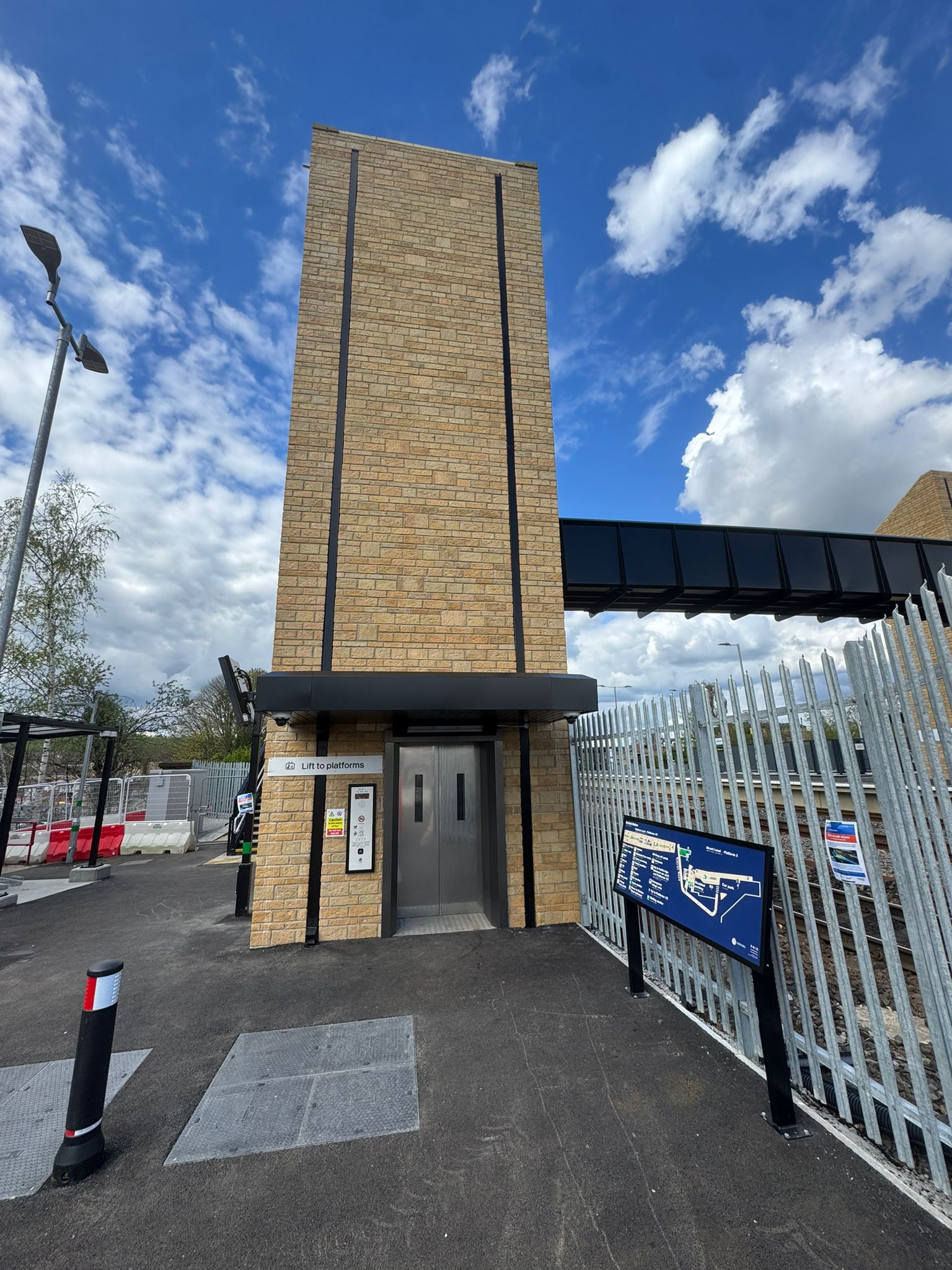 A tall brick clad lift tower stands beside a railway line, with a stainless steel lift entrance at ground level and an elevated footbridge extending from the structure. A station map and information board are positioned nearby, while security fencing runs alongside the tracks. The scene is set beneath a bright blue sky with large white clouds.