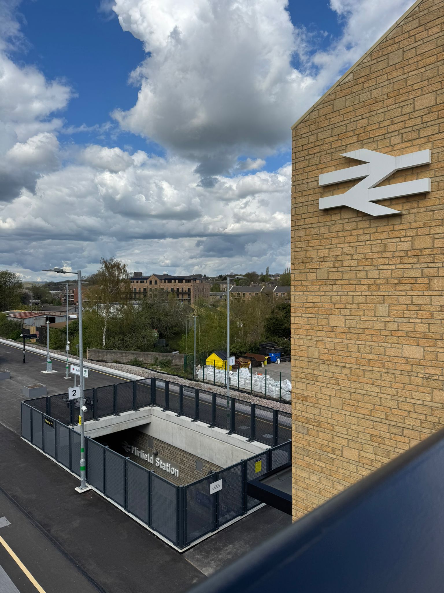Elevated view across Mirfield Station showing platform areas, safety fencing, and the station entrance below. A brick clad tower displaying a large white rail symbol stands in the foreground, while the station sign is visible within the recessed entrance area. Trees, nearby buildings, and dramatic cloud formations fill the background beneath a bright blue sky.