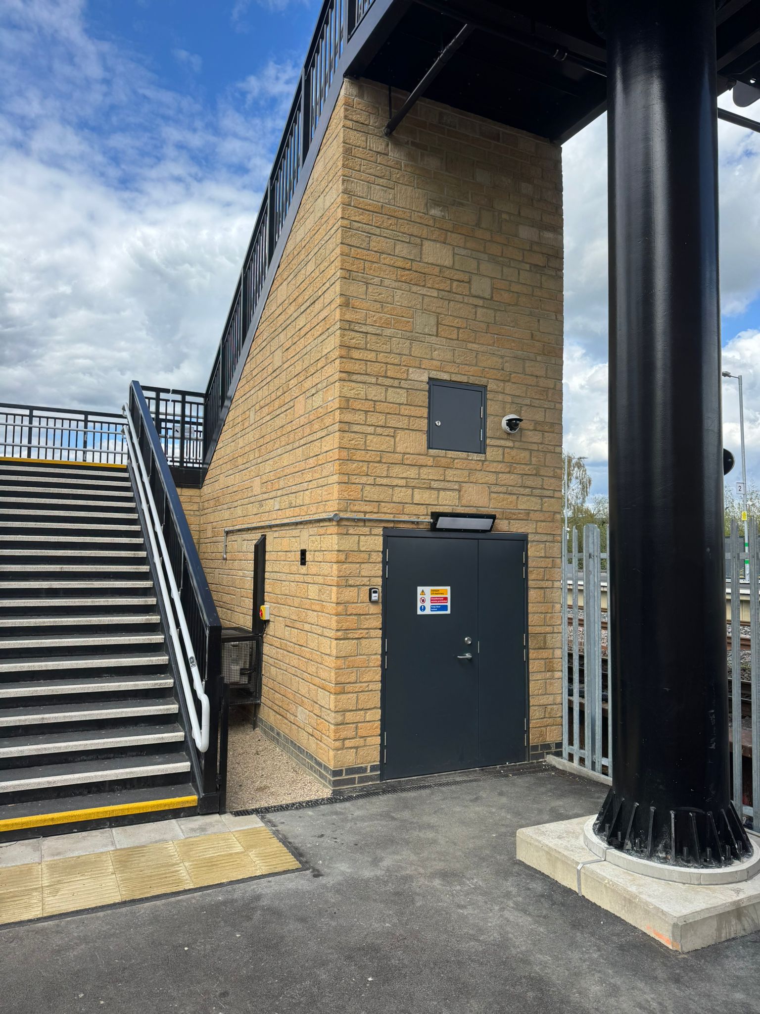 A staircase with black metal railings rises alongside a brick clad station structure. The image shows construction details including access doors, security cameras, tactile paving at the foot of the stairs, and a large steel support column for the overhead footbridge. Railway tracks and fencing are visible in the background beneath a partly cloudy sky.