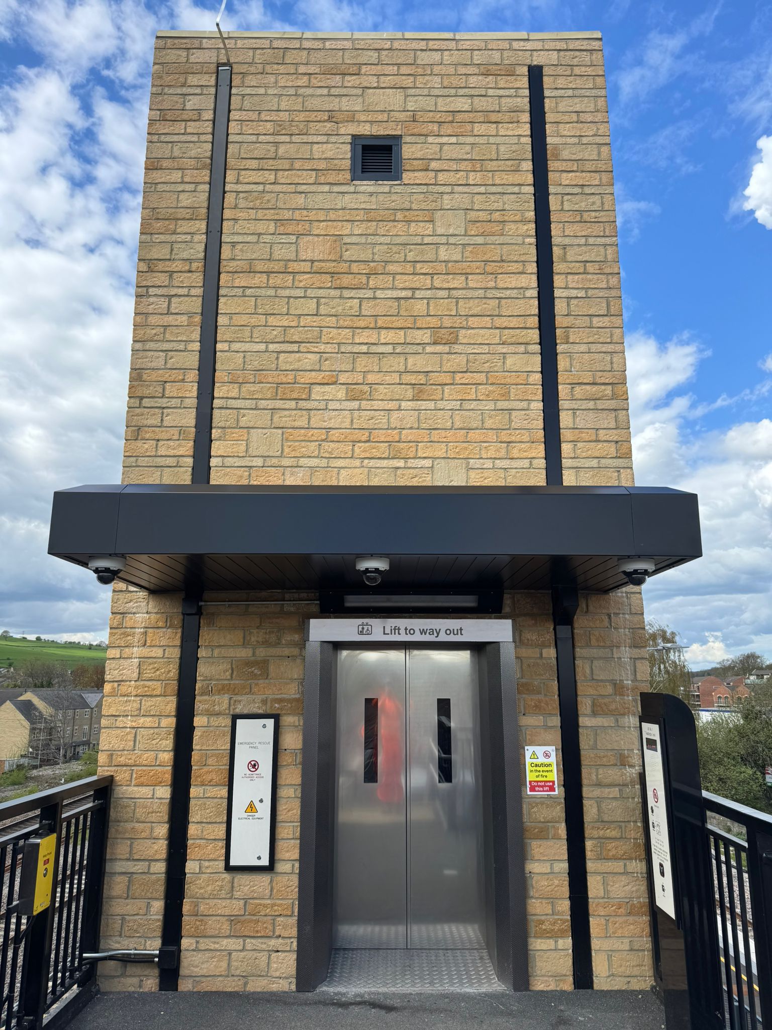 Front view of a brick clad lift tower on an elevated railway platform. Stainless steel lift doors sit beneath a dark canopy with integrated lighting and security cameras. Black metal railings border the platform, while blue sky and scattered clouds provide the backdrop. The image highlights the station's modern accessibility infrastructure and architectural design.