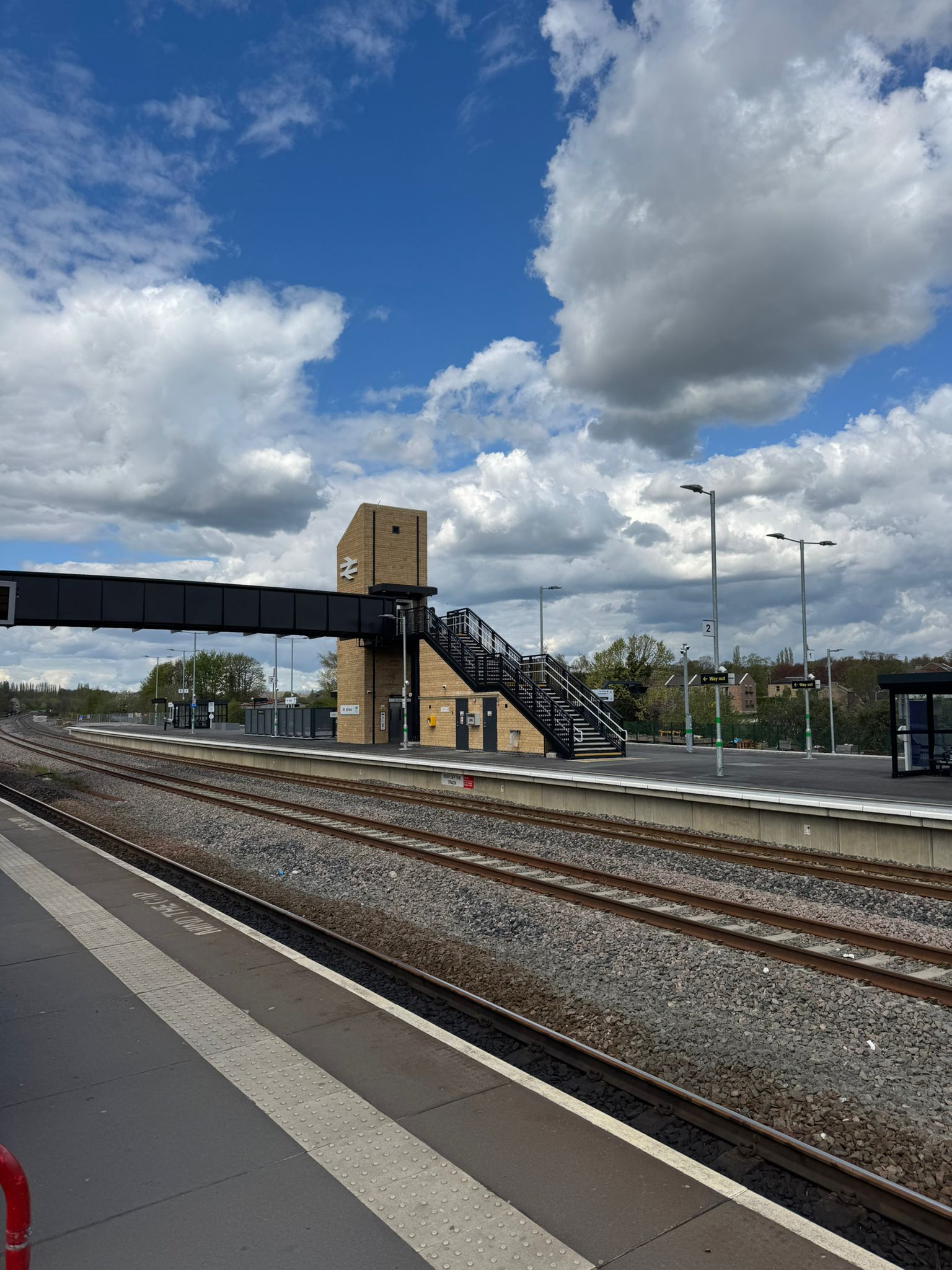 View across multiple railway tracks towards a modern station platform with a brick clad lift tower, staircase, and elevated pedestrian footbridge. Platform shelters, lighting columns, and signage are visible along the platform, while large white clouds fill the blue sky above the station.