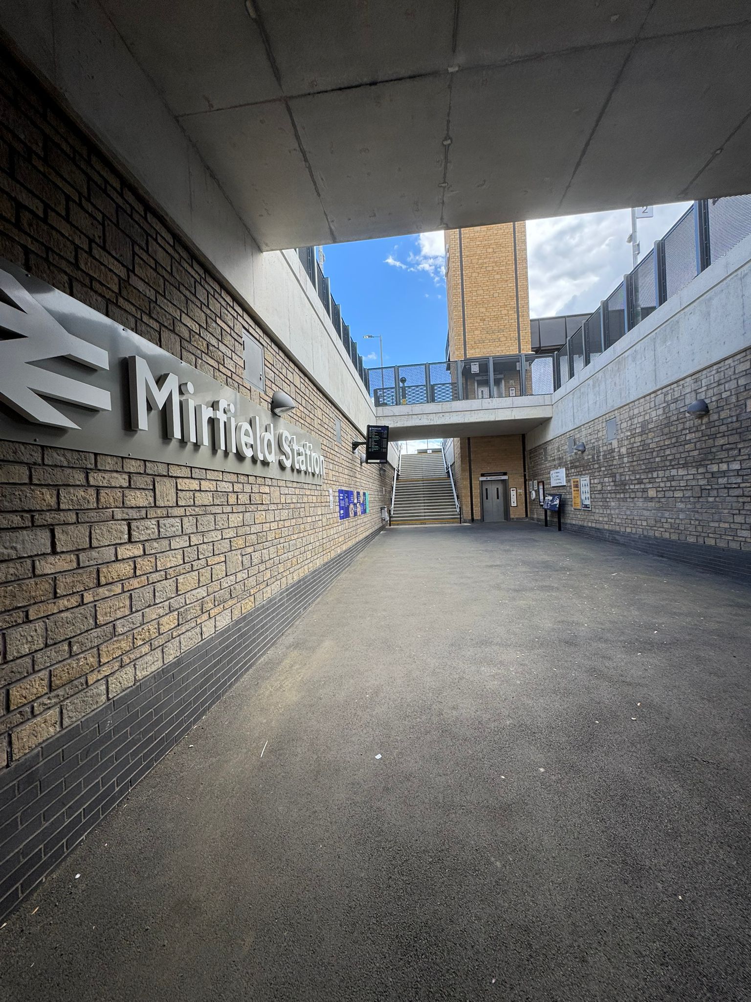 A wide covered station entrance at Mirfield Station with brick walls, concrete structures, and a large station sign mounted along one side. Stairs and a lift provide access to the platforms above, while information boards, lighting, and wayfinding signs line the open pedestrian space. Natural light enters from the open ends of the underpass, highlighting the modern station design.
