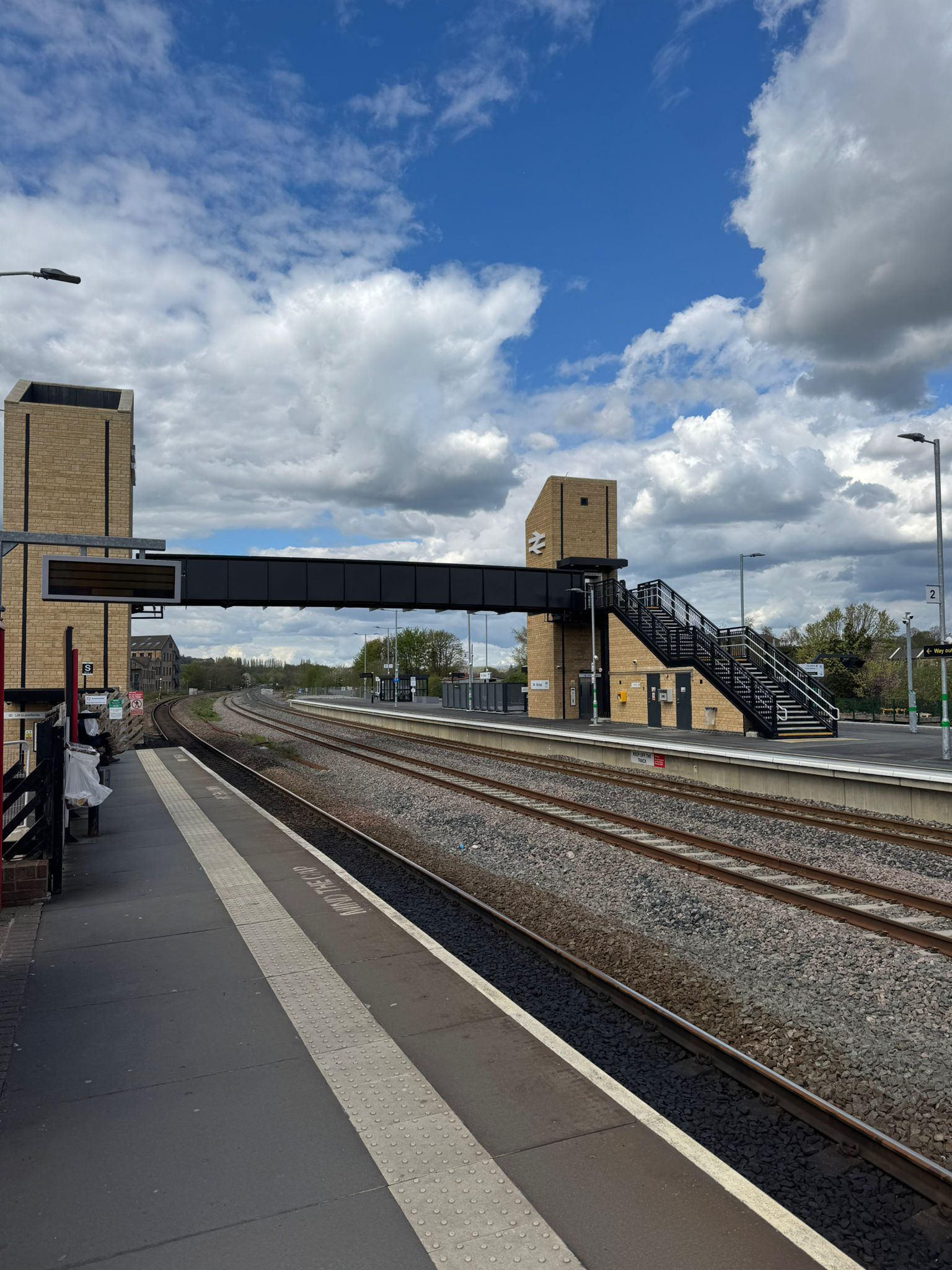 View along a railway platform showing multiple tracks, a modern pedestrian footbridge, and two brick clad lift towers connected by a black enclosed walkway. Platform signage, lighting columns, and tactile paving run alongside the tracks, with dramatic clouds and patches of blue sky overhead.