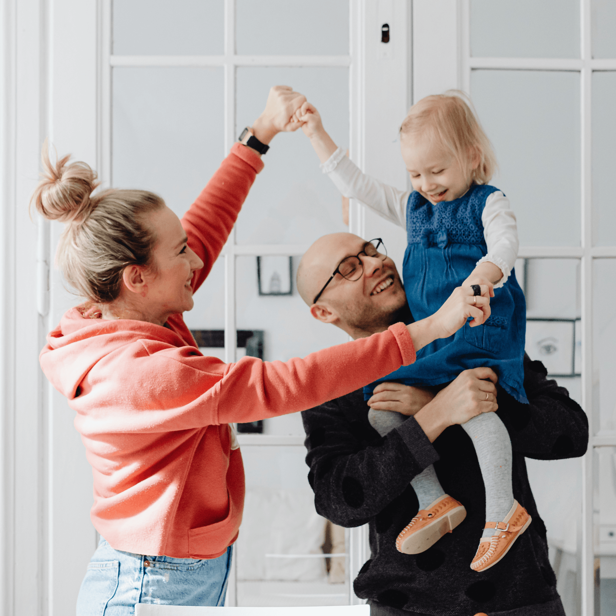 A man carries a child on his shoulders while a woman dances with them, all indoors in front of white French doors.