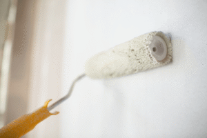 A close-up shot of a paint roller with a yellow handle applying white paint to a wall.