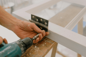 A person uses a drill to attach a metal hinge to a white wooden door frame.